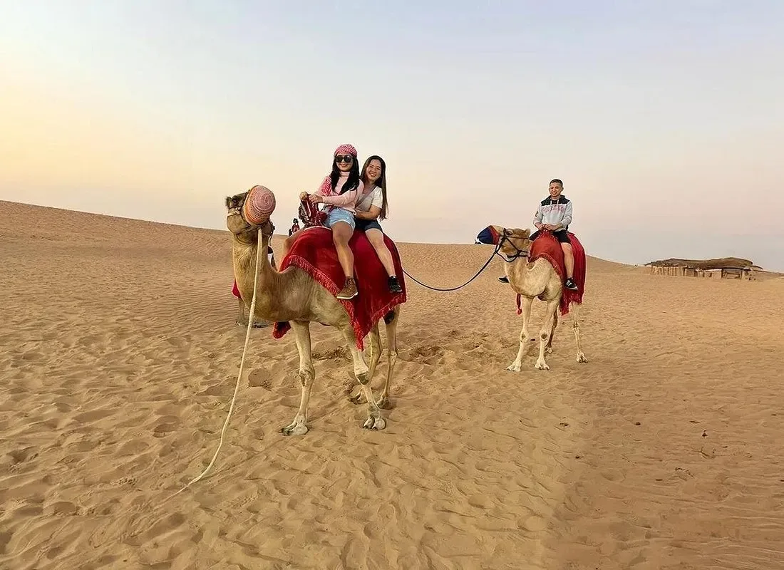 Two guests riding a camel led by a handler across a dune at golden hour on a Dubai desert safari