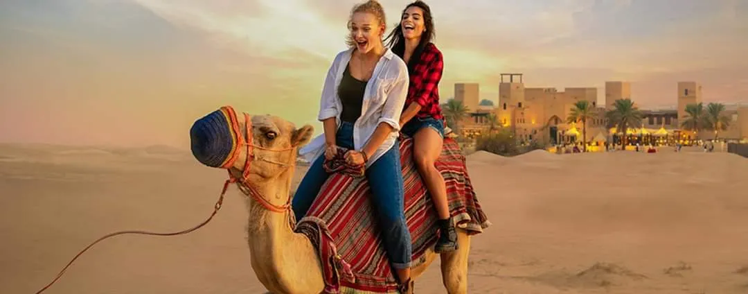 Two guests riding a camel across the dunes with a desert fort and palm trees visible in the distance