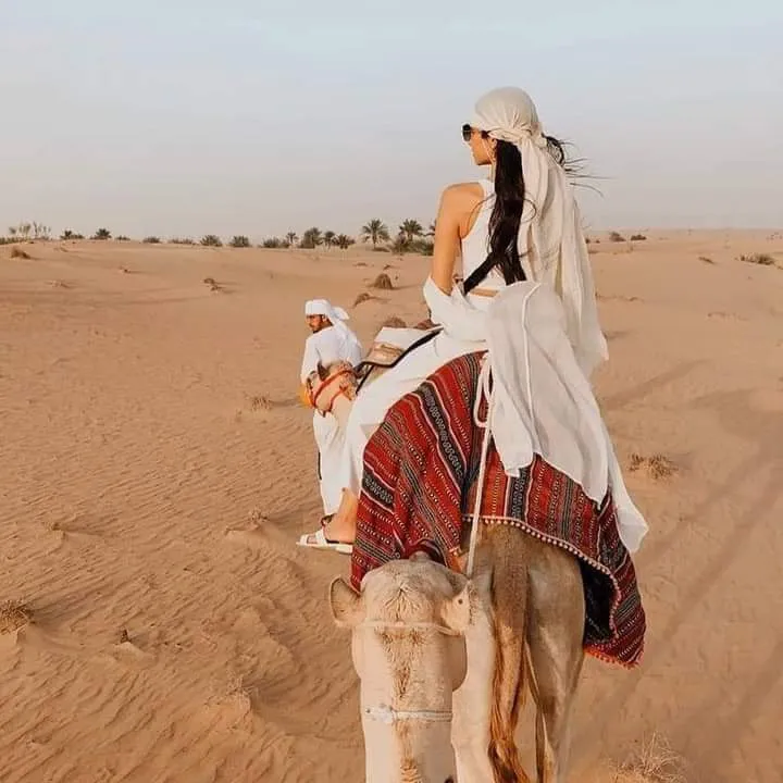 Two guests riding a camel led by a handler along a desert path lined with palm trees in soft morning light