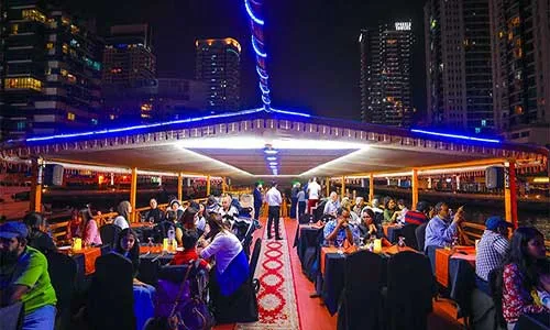 Guests dining on the dhow’s covered upper deck at night with Marina towers behind