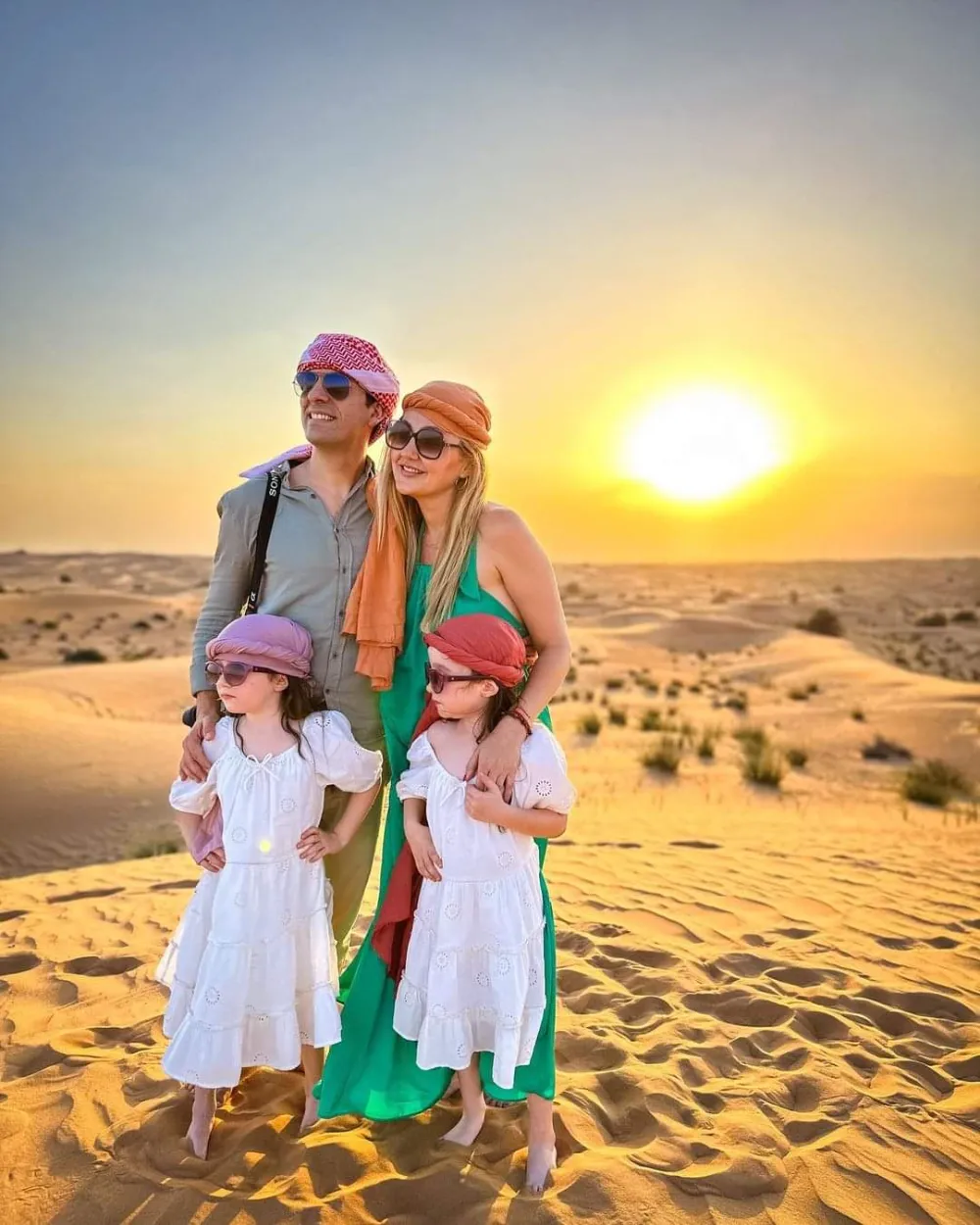 A family enjoying an overnight desert safari in Dubai A family group in traditional red-and-white headscarves posing together on a dune at sunset during a Dubai desert safari