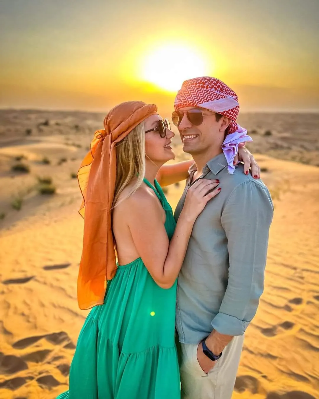 A couple standing close together on a dune with a huge golden sun rising low over the desert horizon